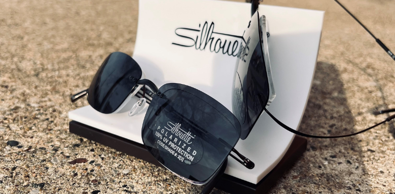 Two pairs of Silhouette eyeglasses with polarized sunglasses clips on a stand outdoors.